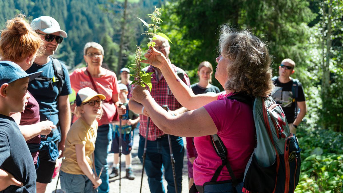 Frau hält Kräuter in der Hand, rund um sie eine Gruppe von Menschen, die ihr zuhören.