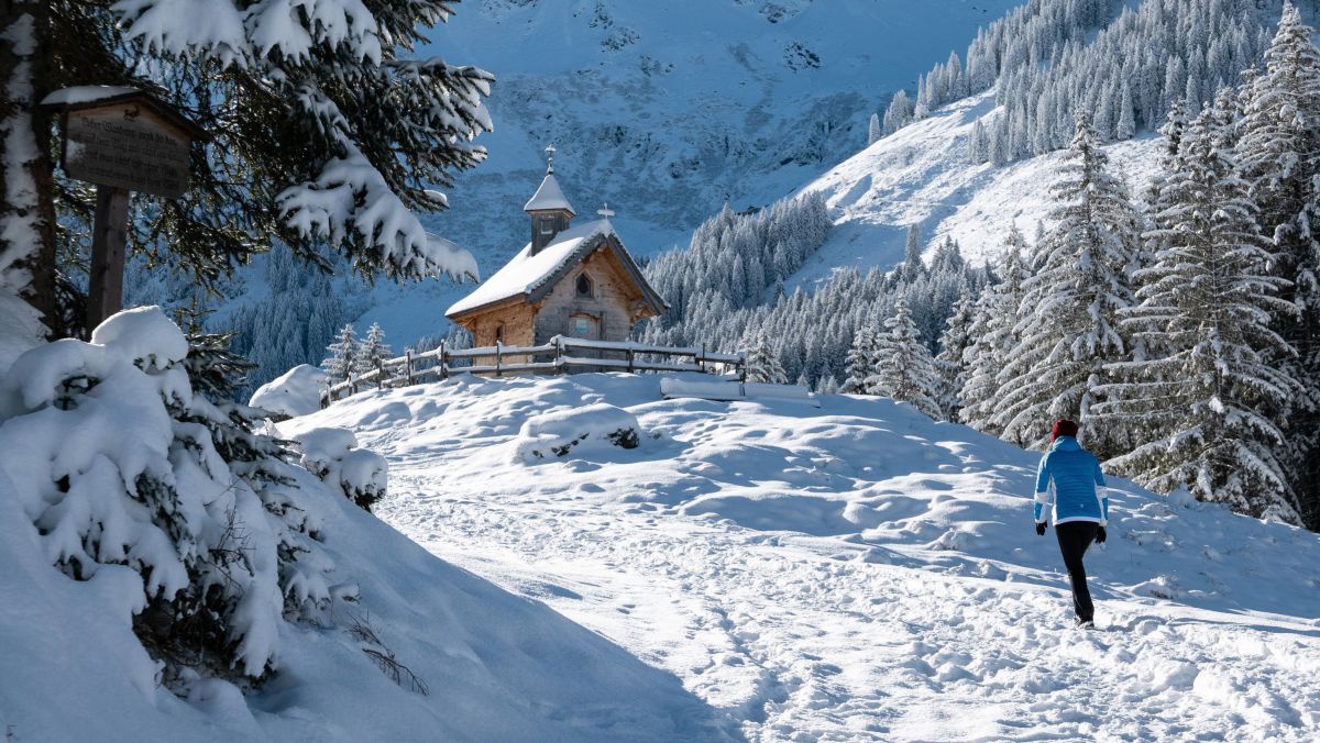 Frau stapft durch den Schnee hoch zu einer Kapelle in einer Waldlichtung.