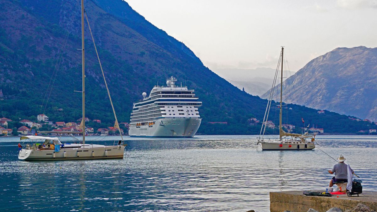 Kreuzfahrtschiff in der Bucht von Kotor, davor 2 Segelschiffe, am Ufer ein Angler.