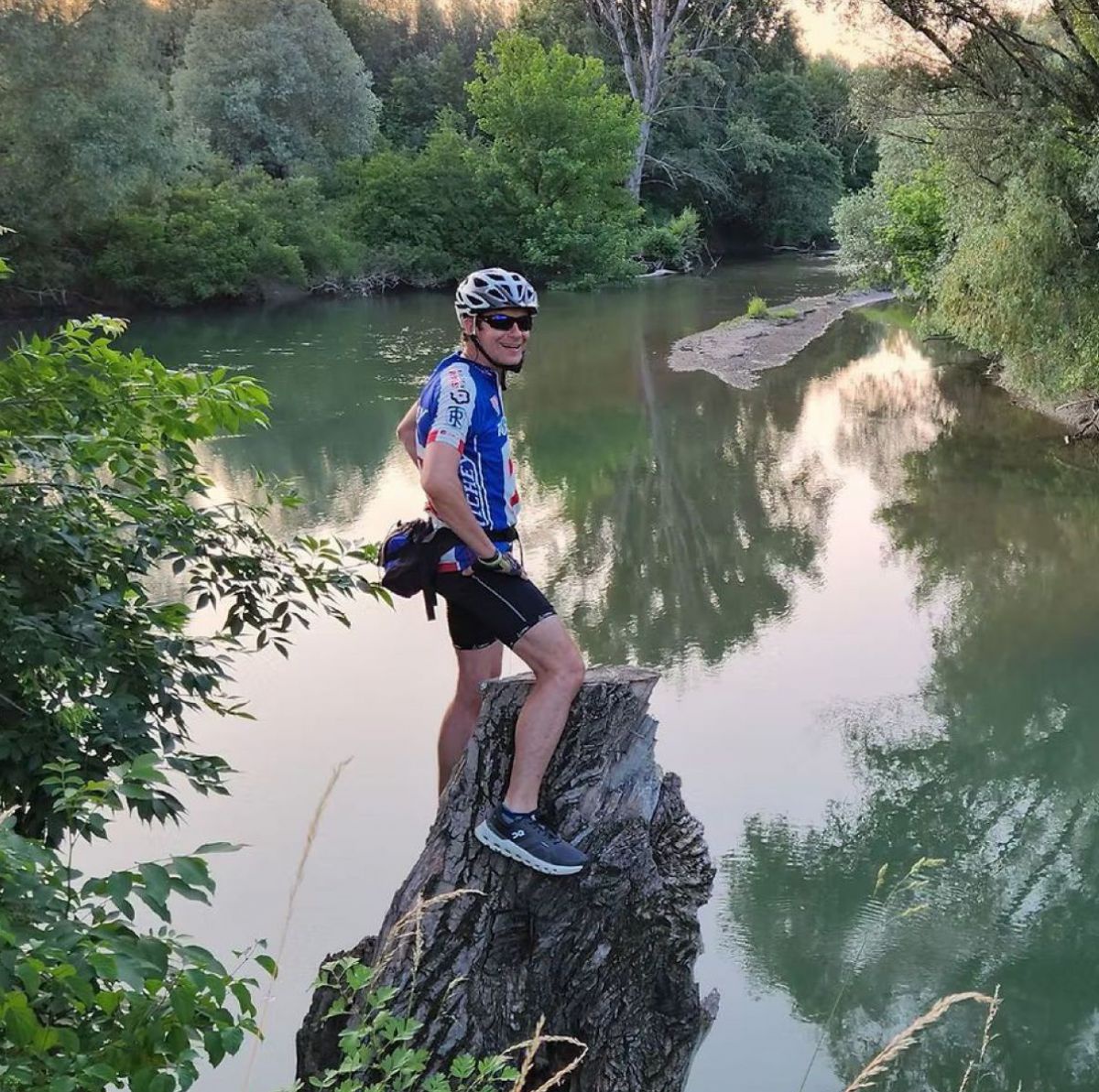 Thomas Schuh steht in Fahrraddress auf einem Baumstumpf am Ufer eines Flusses.