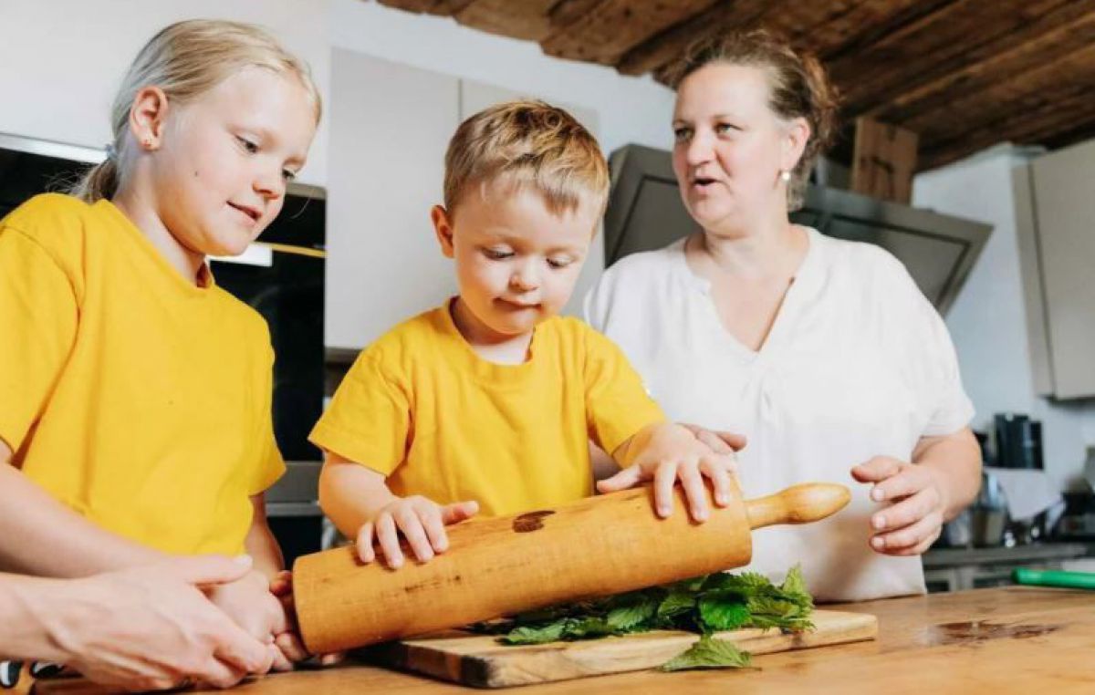 Frau mit 2 Kindern an einem Tisch, der Bub hat ein Nudelholz, das er über einen Buschen Brennesseln rollt.