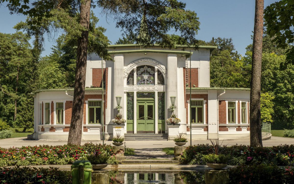 Jugendstilgebäude (weiß mit roten Ziegelsteinen), grünes Einhangstor, umgeben von Bäumen. Im Vordergrund ein Teich.