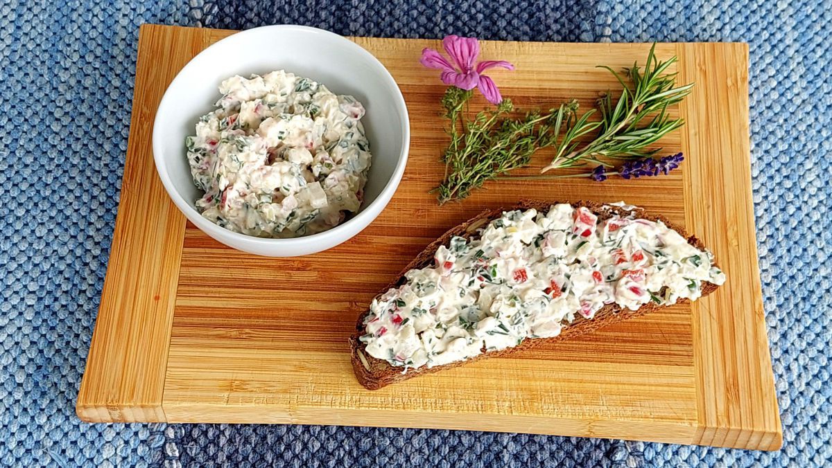 Auf einem Brett liegt ein Brot mit Kräuteraufstrich, daneben eine Schüssel mit diesem Aufstrich und einige Kräurer.