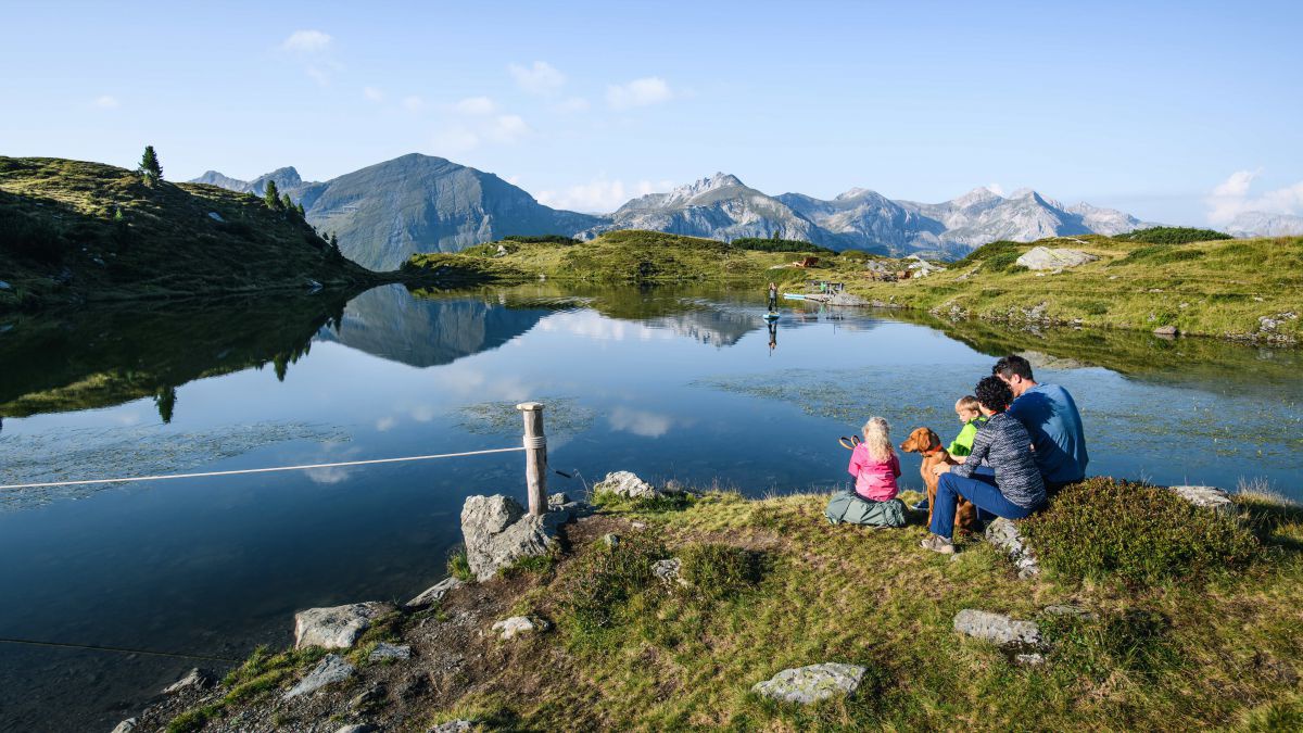 Familie mit 2 Kindern und Hund sitzt am Ufer eines Bergsees. Am See ein Stand Up Paddler. Im Hintergrund Bergpanorama.
