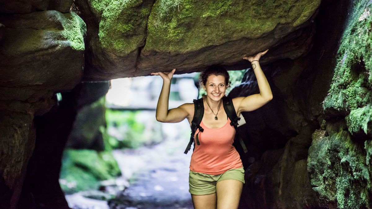 Frau geht unter einem Durchlass aus Felsen, hält die Hände nach oben, was den Anschein erweckt als würde sie den riesigen Stein über ihr halten.
