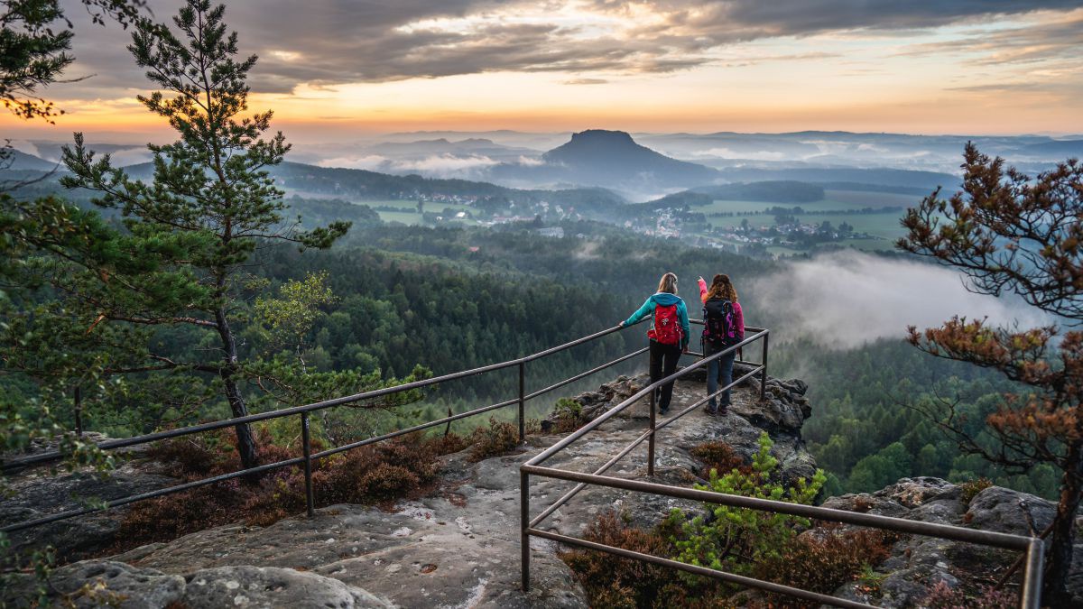 2 Personen stehen in der Morgendännerung auf einer Aussichtsplattform. Nebel hängt in den Wäldern darunter. Der Blick geht weit hinein ins Elbtal. In der Mitte ein Tafelberg.
