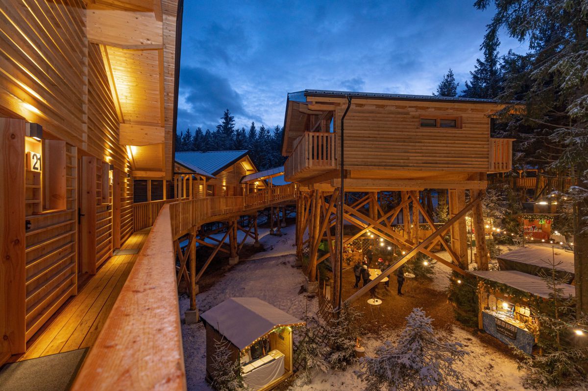 Blick auf die Holzgebäude des Baumhotels im Winter: Die Häuser sind angenehm beleuchtet, auf den Dächern, den Bäumen und im Hof liegt etwas Schnee. Menschen stehen zwischen Hüttchen und Ständen bei Feuerplätzen, darüber hängen Lichterketten.