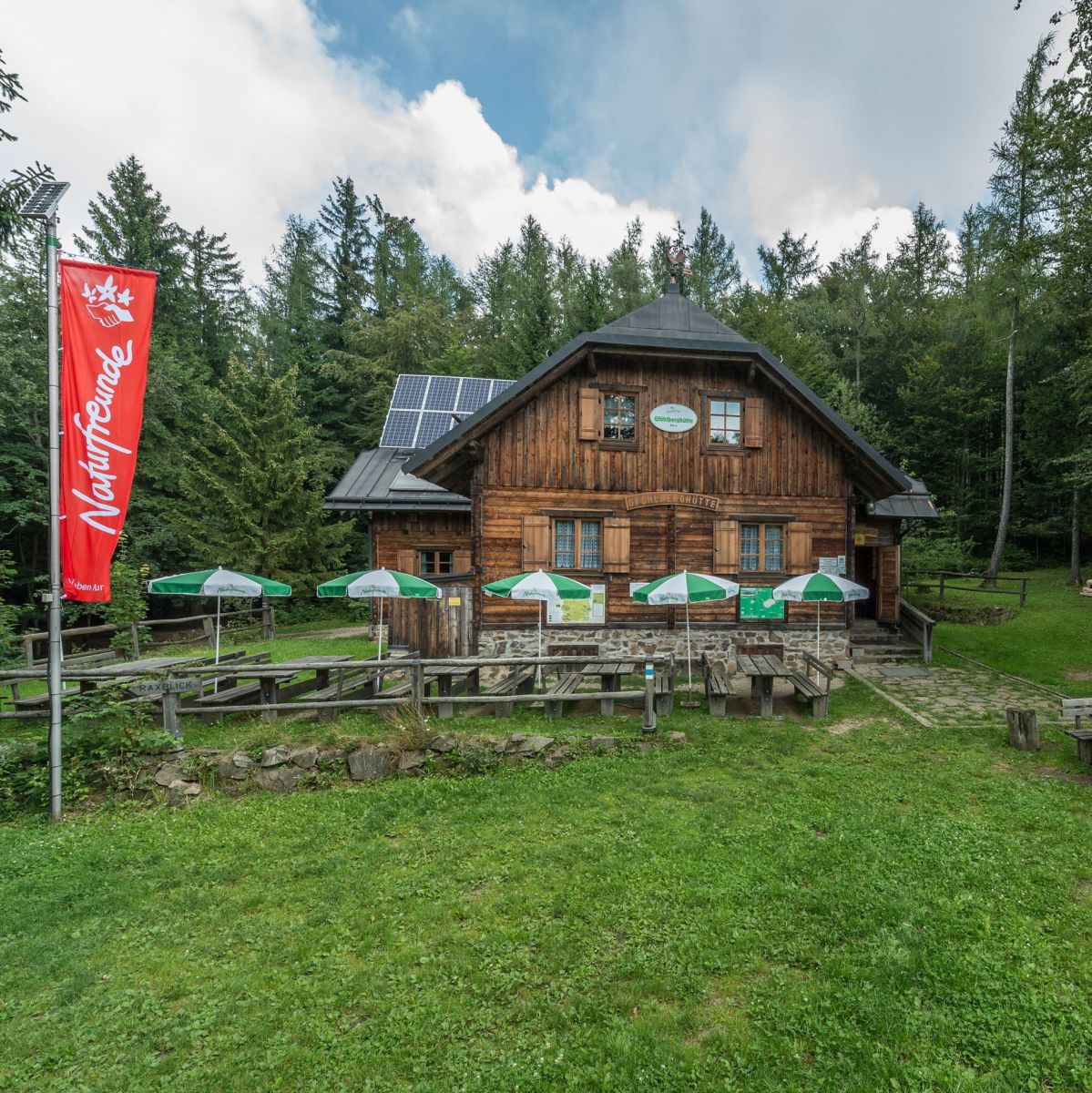 Naturfreundehaus aus Holz, Terasse mit grün-weißen Sonnenschirmen, vor der Hütte eine rote Naturfreunde-Fahne.