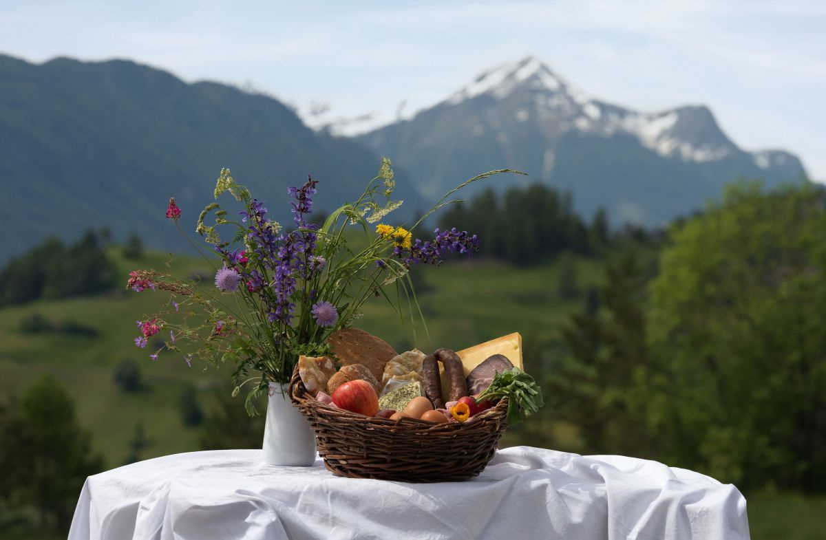Tisch mit weißem Tischtuch im Grünen, darauf ein Korb mit Obst, Fleisch, Brot usw, daneben in einer Vase ein Strauß Wiesenblumen. Dahinter ein zum Teil schneebedecktes Bergmassiv.