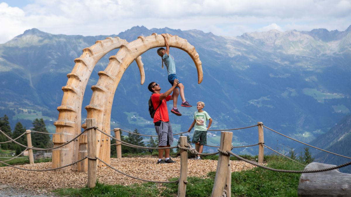 Zwei riesige Mammutzähne aus Holz. Mädchen klettert rauf, Mann sichert sie dabei.