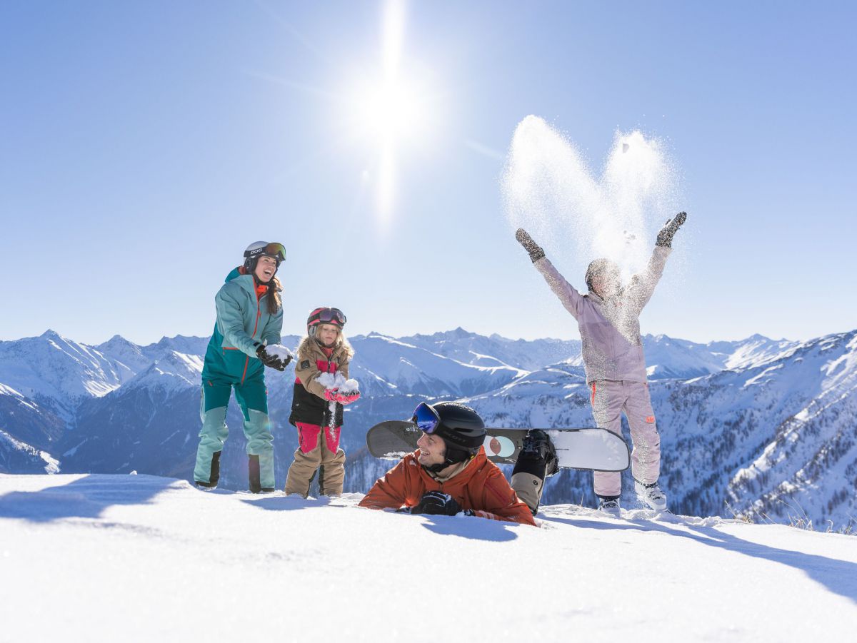 Familie tobt im Schnee herum. Im Hintergrund ein Gebirgsmassiv und die Sonne scheint dazu.