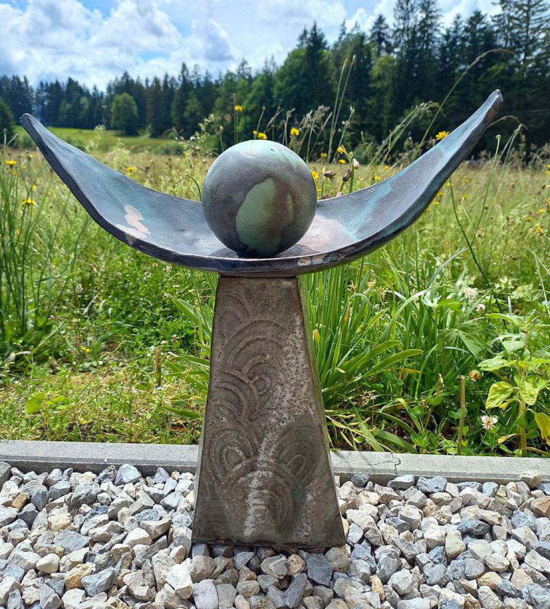 Skulptur aus konischem Sockel, Metallschale und einer Kugel darin ergibt die Form einer Figur mit ausgebreiteten Armen.