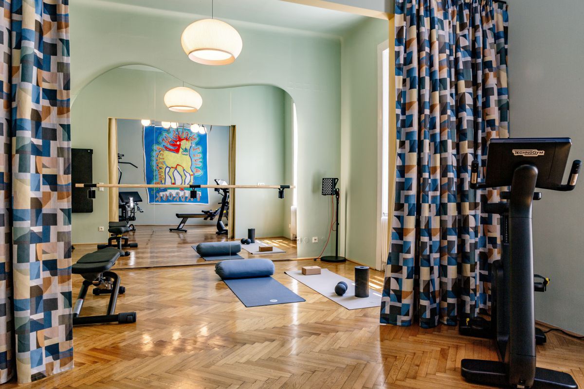 Fitnessgeräte, Bodenmatten, Vorhang mit geometrischen Mustern. Hinten im Raum ein Spiehel, in dem sich ein buntes Bild spiegelt.