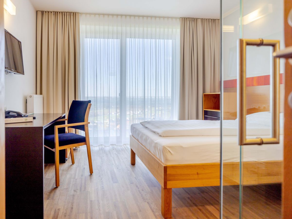 Zimmer mit Bett, Schreibtisch, Sessel. Hinter dem zugezogenen Vorhang führt der Blick über den etwas tiefer liegenden Ort Schlierbach.