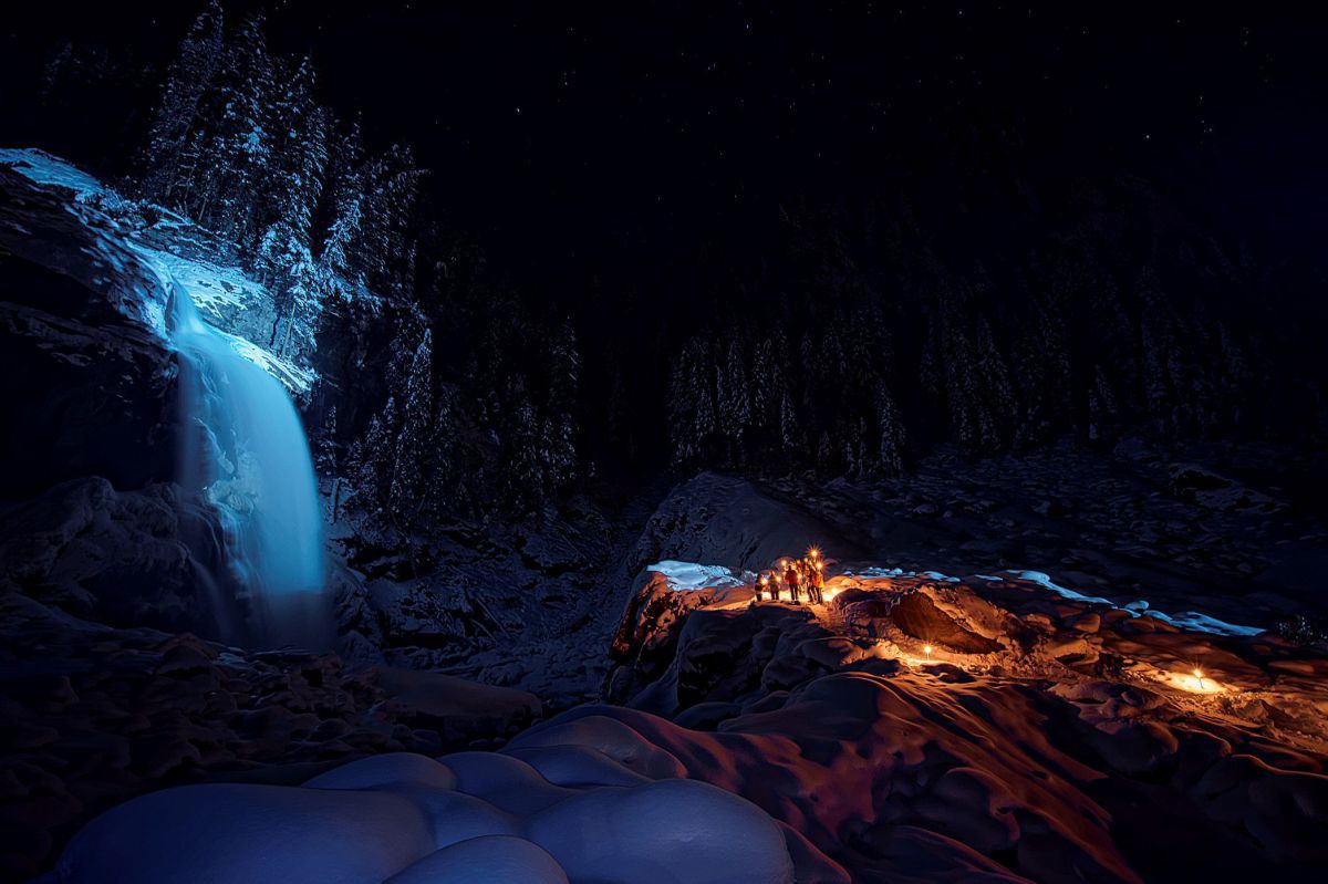 Eine Gruppe von Menschen mit Lampen bei Nacht auf einem Felsen neben einem beleuchteten Wasserfall in einer verschneiten Landschaft.