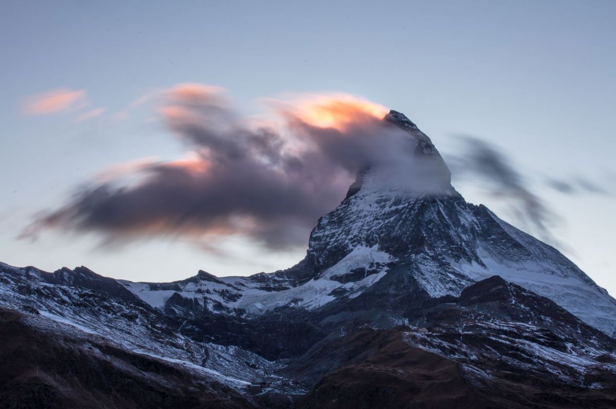 Ein schneebedeckter, sehr spitzer, hornförmiger Berggipfel, halb von Wolken verdeckt, etwas Sonnenlicht glüht hinter dem Gipfel hervor.