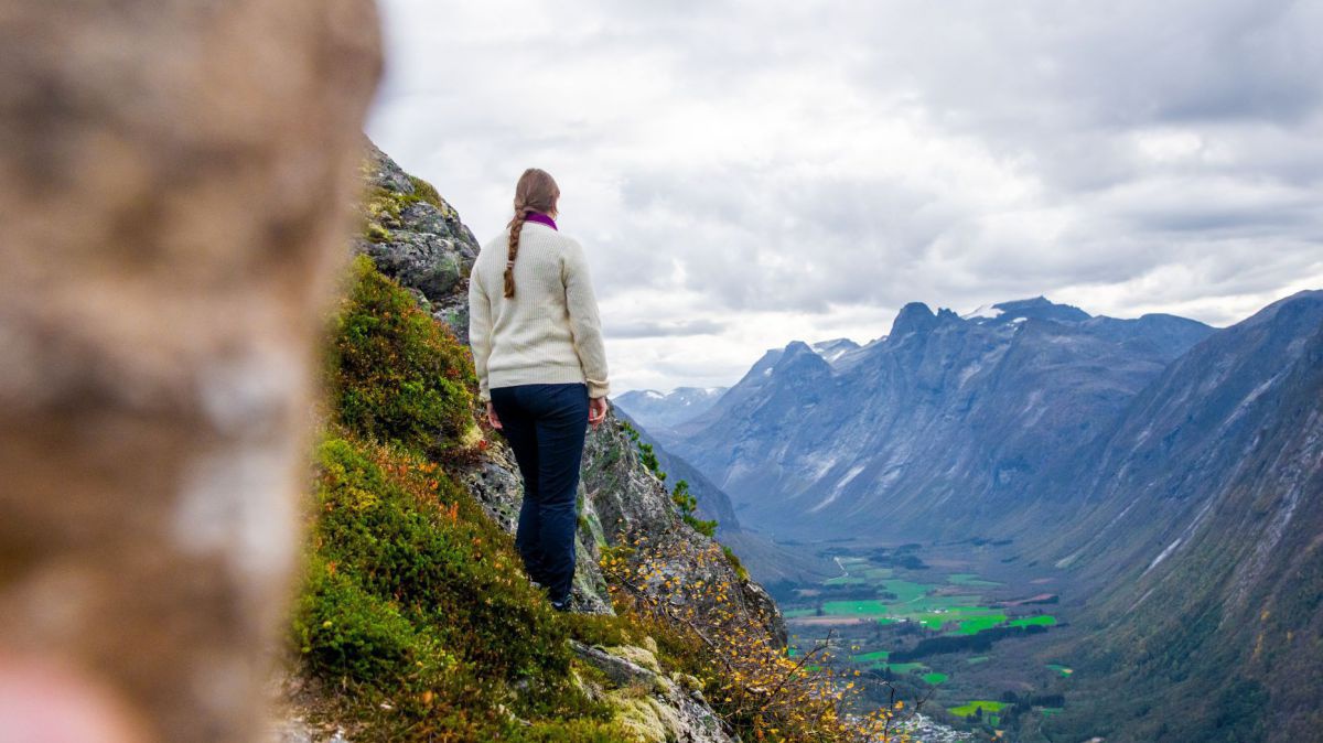 Eine Person mit geflochtenem Haar und weißem Pullover steht auf einem bewachsenen Felsen und blickt auf ein Tal und dahinter eine steil aufstrebende Felswand der gegenüberliegenden Berge.