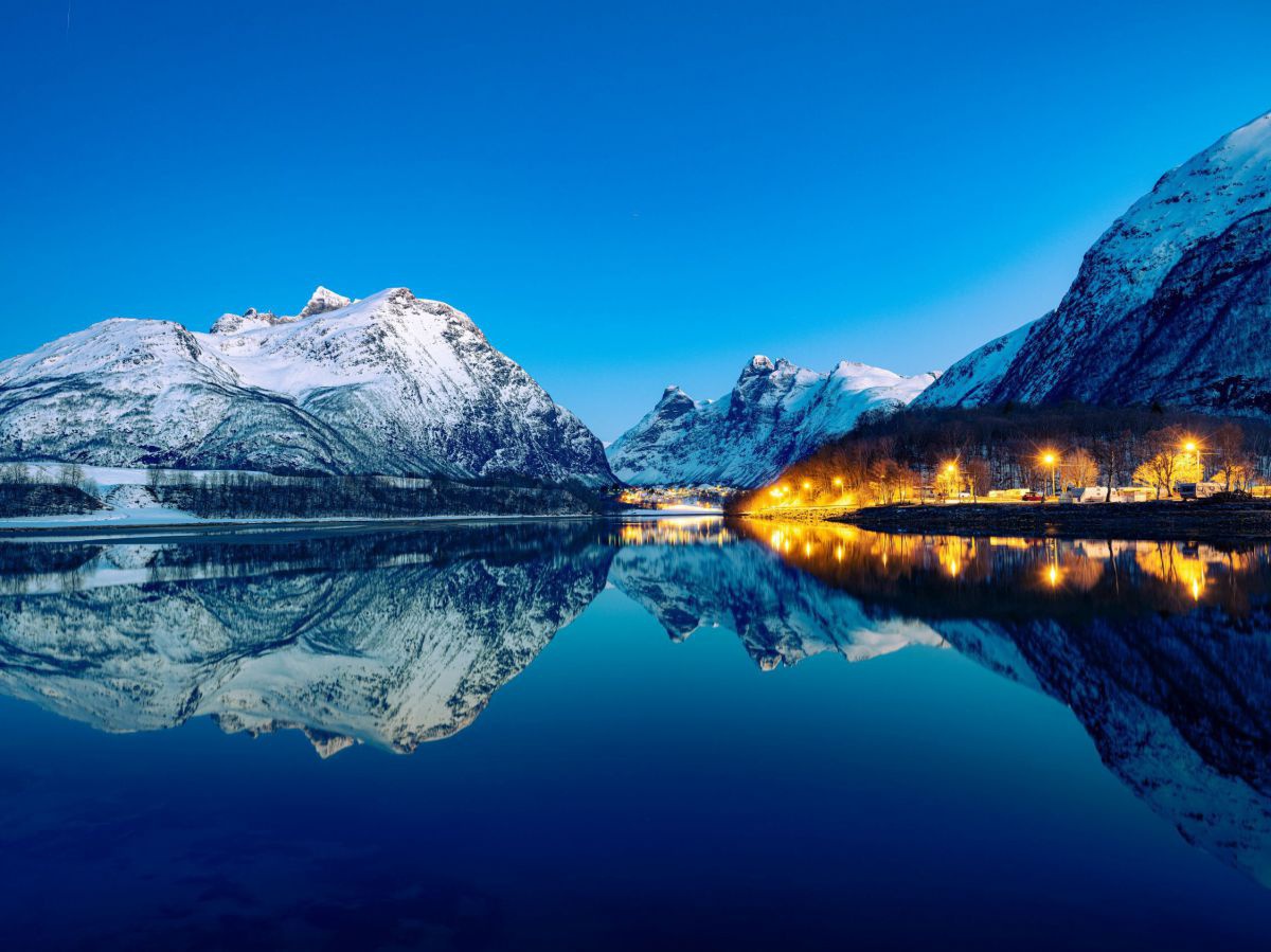 Schneebedeckte Berge spiegeln sich bei Dämmerung in einem ruhigen See während am Ufer die Straßenlaternen an der Promenade warm leuchten.