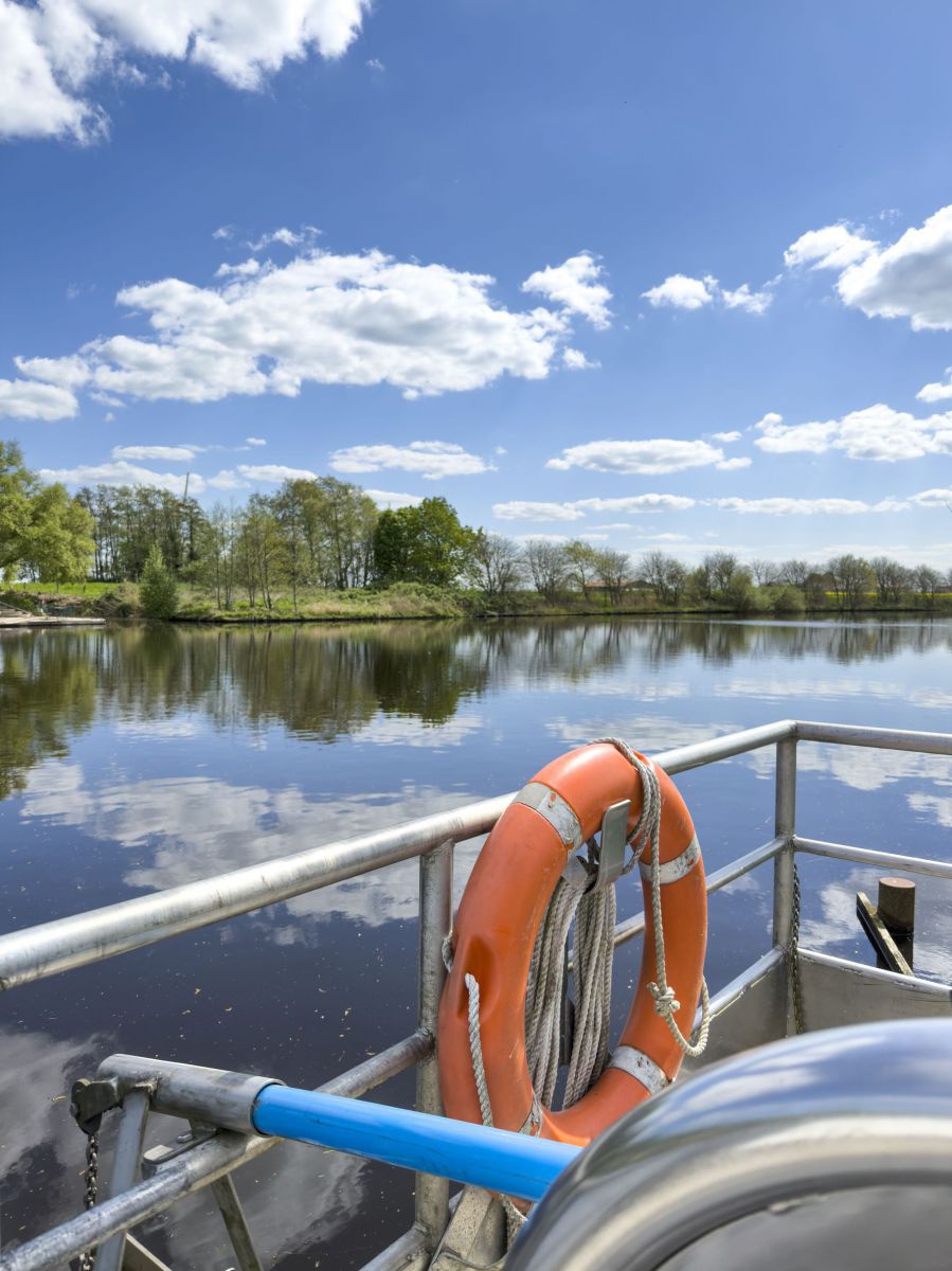 Ein orangefarbener Rettungsring hängt an der Reling eines Bootes mit Blick auf das ruhige Wasser eines Sees in dem sich der blaue, leicht bewölkte Himmel und die Bäume am anderen Ufer spiegeln.