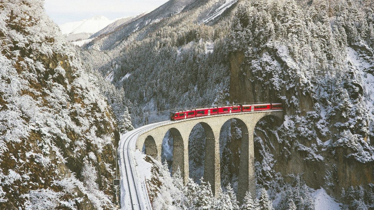 Blick auf einen roten Zug, der aus einem Tunnel fährt und dabei ist, die Schlucht zwischen zwei verschneiten, bewaldeten Felsen auf einem Viadukt zu überqueren.