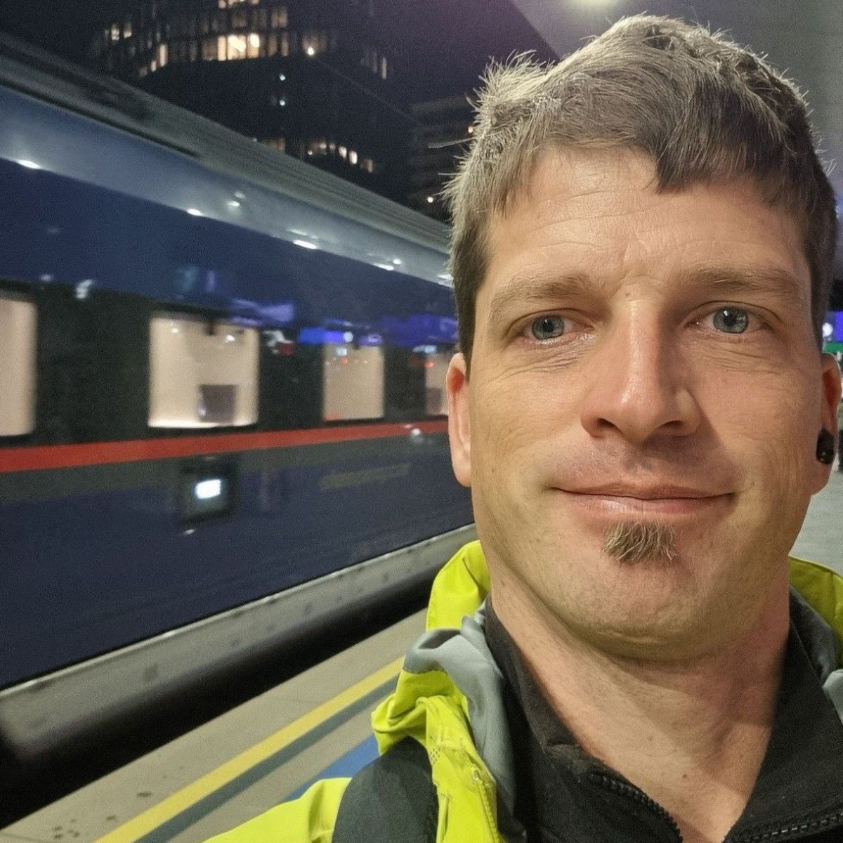 Eine Person mit kurzem Haar und gelber Jacke macht ein Selfie auf einem Bahnsteig während im Hintergrund ein Zug vorbeifährt.