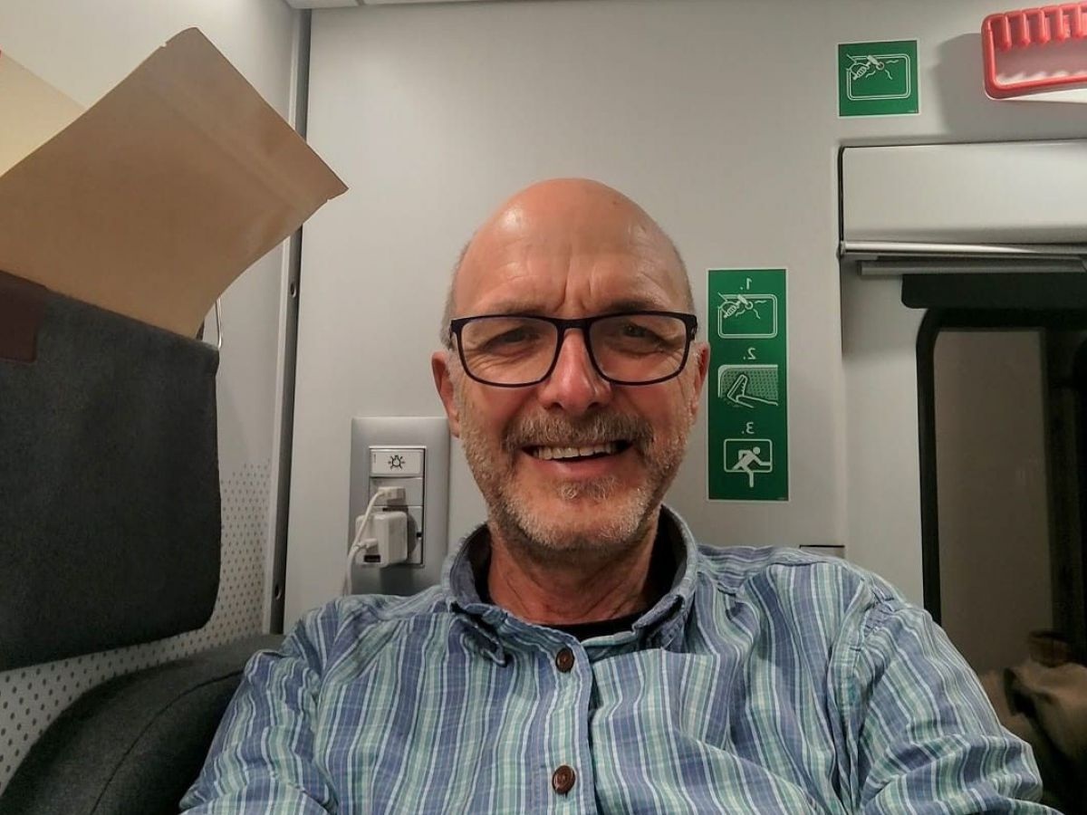 Eine lächelnde Person mit Brille und kariertem Hemd macht ein Selfie im Inneren eines Zuges. Im Hintergrund auf der Wand sieht man ein grünes Notausgangsschild und eine Steckdose.