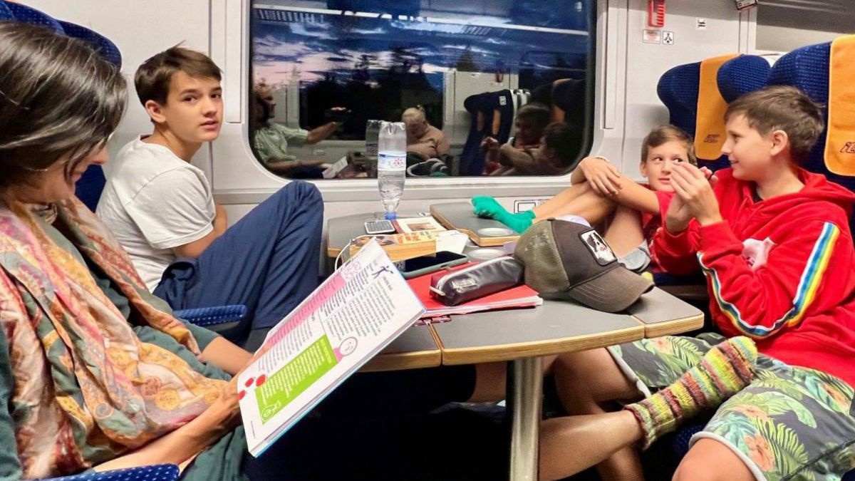 Vier Personen sitzen in einem Zugabteil: die Frau liest und die drei Kinder unterhalten sich angeregt während auf dem Tisch zwischen ihnen Gegenstände wie Schreibzeug, Bücher, Hefte, Trinkflasche und eine Kappe liegen.