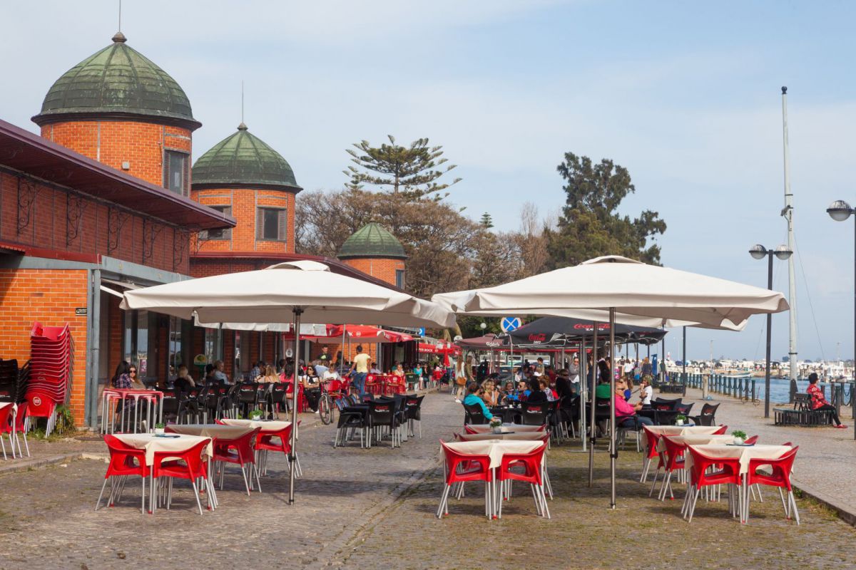 Eine belebte Aussenterrasse mit roten Stühlen und großen, weißen Sonnenschirmen vor einem ziegelroten Gebäude mit runden Türmen und grünen Haubendächern an einem sonnigen Tag am Wasser.