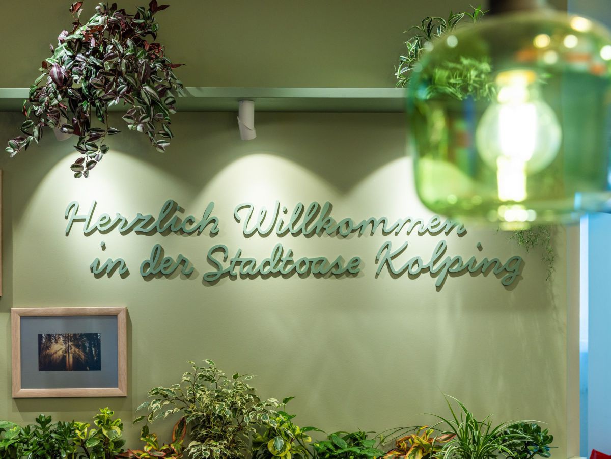 Grüner Schriftzug an einer Innenwand: Herzlich Willkommen in der Stadtoase Kolping, dazu grüne Pflanzen.