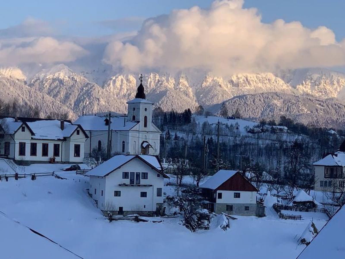 Verschneites Dorf, dahinter eine mächtige Gebirgskette in der Abendsonne.