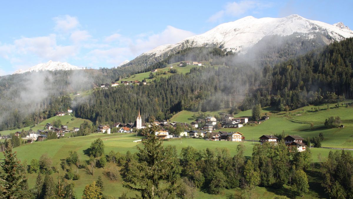 Blick auf eine kleine Gemeinde mit Kirchturm im Zentrum. Daginter ein mächtiger verschneiter Bergrücken.