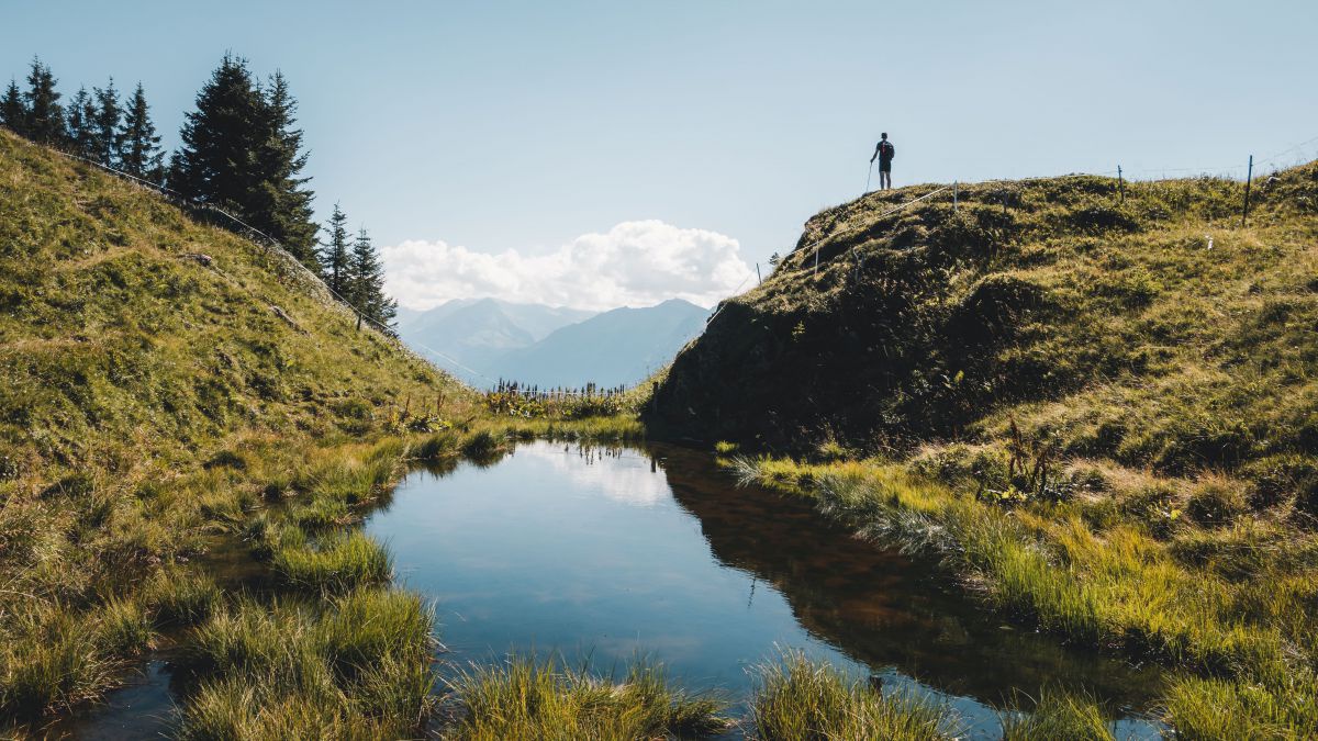 Kleiner Bergsee, Wanderer steht etwas oberhalb und blickt hinaus in ein Bergpanorama.