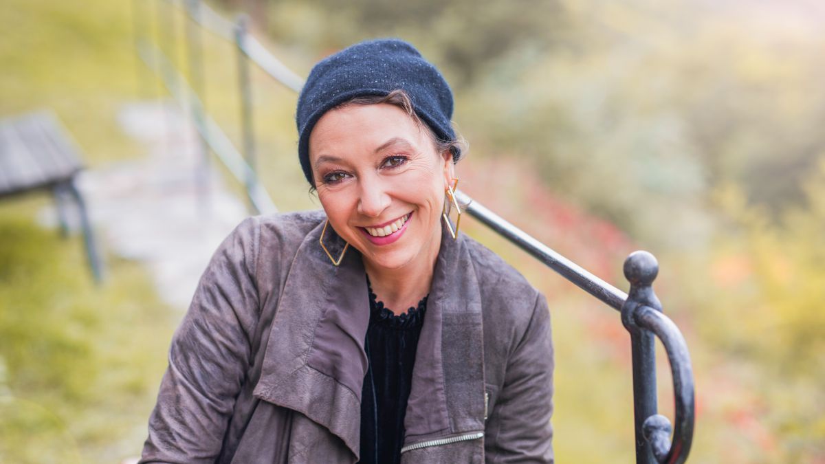 Ursula Strauss mit blauer Wollmütze in der Landschaft, hinter ihr ein eisernes Geländer und eine Sitzbank.