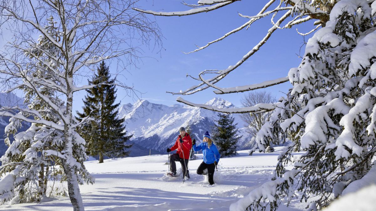 Mann und Frau stapfeen durch eine tief verschneite Landschaft, dahinter ein mächtiger Gebirgszug.