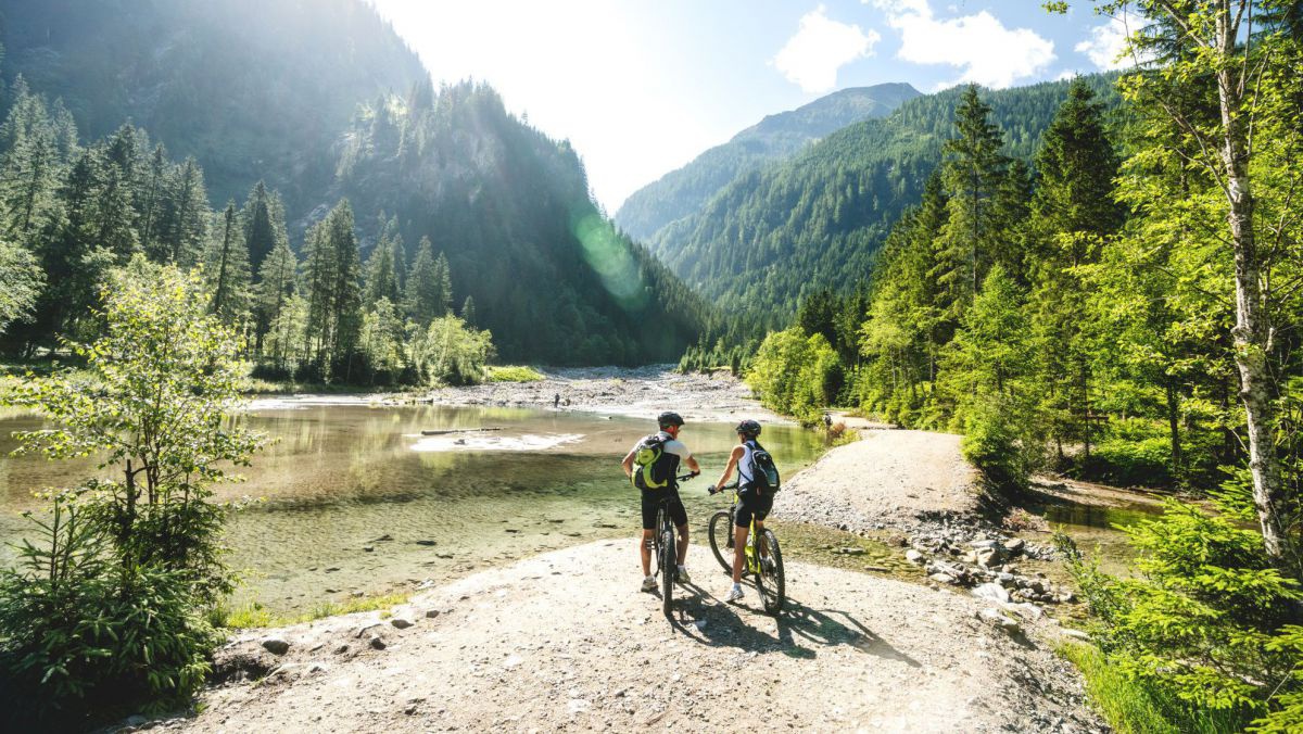 2 Radfahrer stehen vor einem seichten Bergsee, der Radweg durchquert einen Abfluss des Sees.