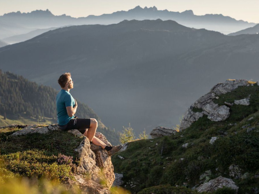 Mann sitzt meditierend auf einem Felsvorsprung. Im Hintergrund Bergpanorama.