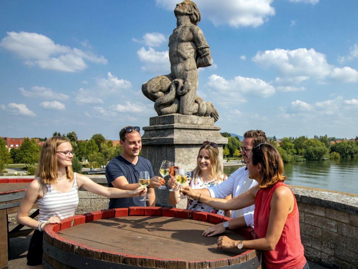 Gruppe von fünf Personen stehen um ein Weinfass und prosten sich zu. Im Hintergrund eine steinerne Statue und der Blick aif einen Fluss.