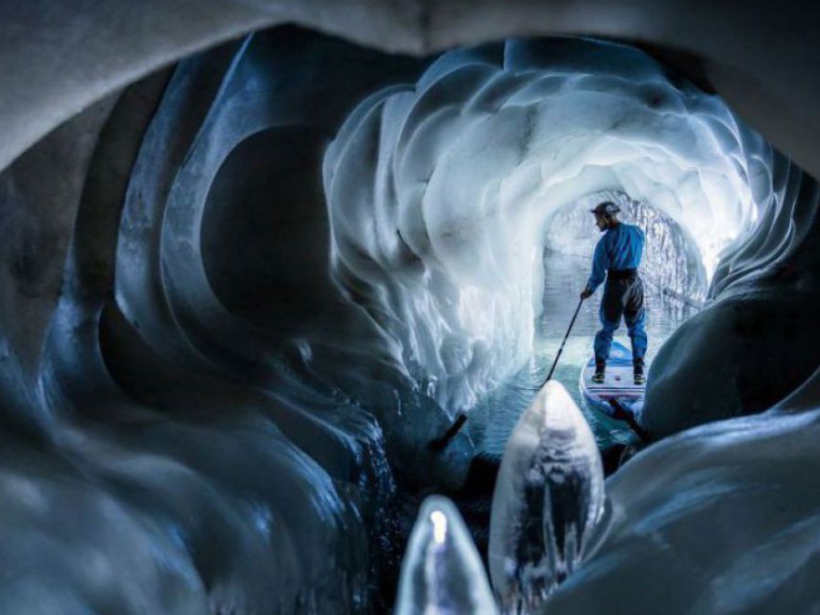 Im Inneren einer Eishöhle, durch deren Gänge sich ein Gletschersee verzweigt: Die eisigen Wände schimmern bläulich, ein Mann erkundet auf einem Stand-Up Paddle die Höhle.