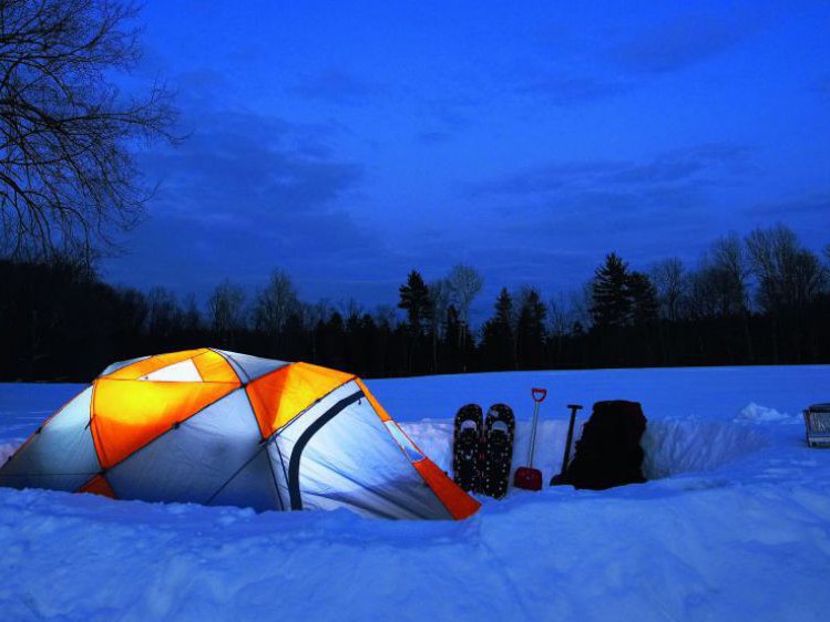 Ein von innen beleuchtetes Zelt auf einer verschneiten Wiese am Waldrand. Der Himmel ist dunkelblau, das Zelt leuchtet hellblau und orange. Neben dem Zelt stehen Schneeschuhe, Rucksack, Schneeschaufeln.