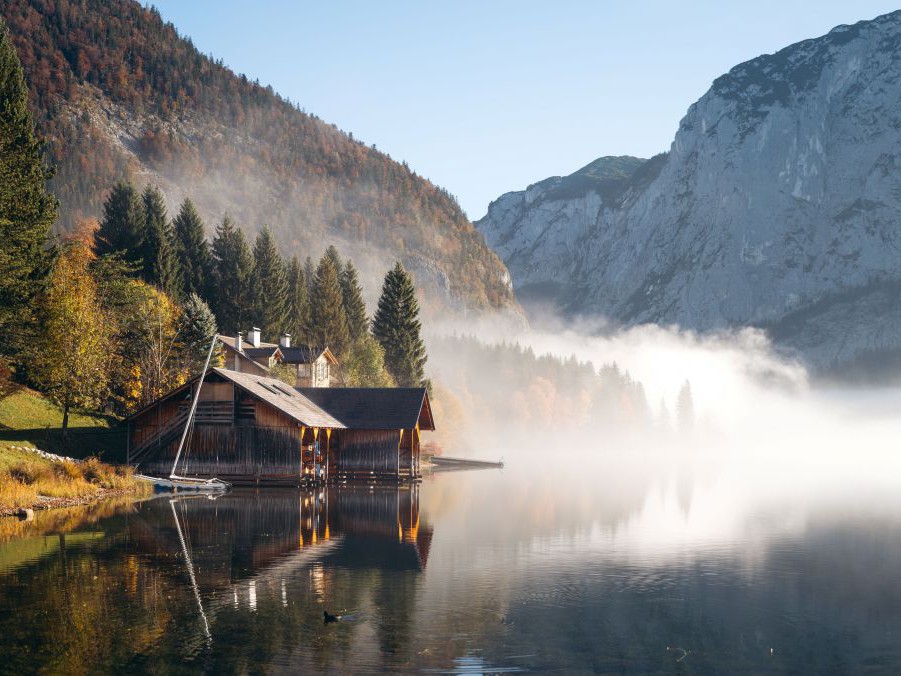 See, am Ufer ein Haus, Bäume sind herbstlich gefärbt, am Wasser liegt Nebel.
