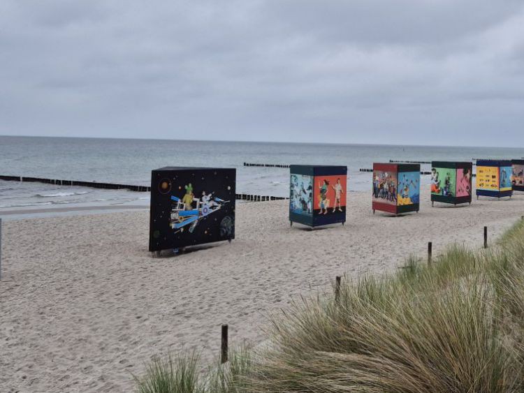 Ein Sandstrand unter tief bewölktem Himmel, ein Teil ist hoch mit Riedgras bewachsen. Den Strand entlang ist eine Kunstausstelllung mit Bildern aufgebaut.