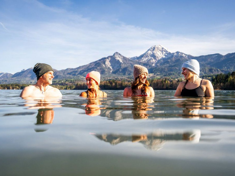4 Personen stehen im Wasser, alle haben eine Haube auf, hinter dem See eine Bergkulisse.