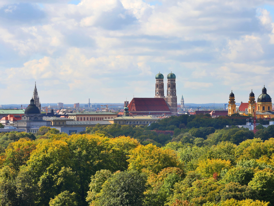 Häuserdächer und Kirchtürme zwischen Baumkronen und sonnig bewölktem Himmel.