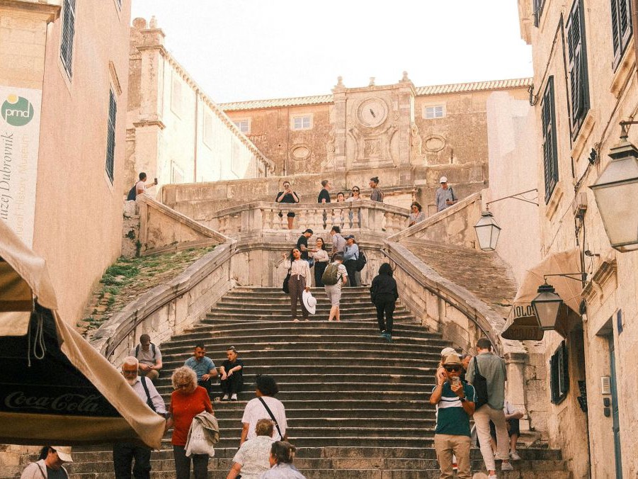 Zwischen hohen Stadhäusern mit dunkelgrünen Fensterläden führt eine breite, lange, teilweise schlängelnde, steinerne Treppe eine Anhöhe hinauf. Ganz oben steht ein dominantes Gemäuer mit großer Uhr über dem Eingang.