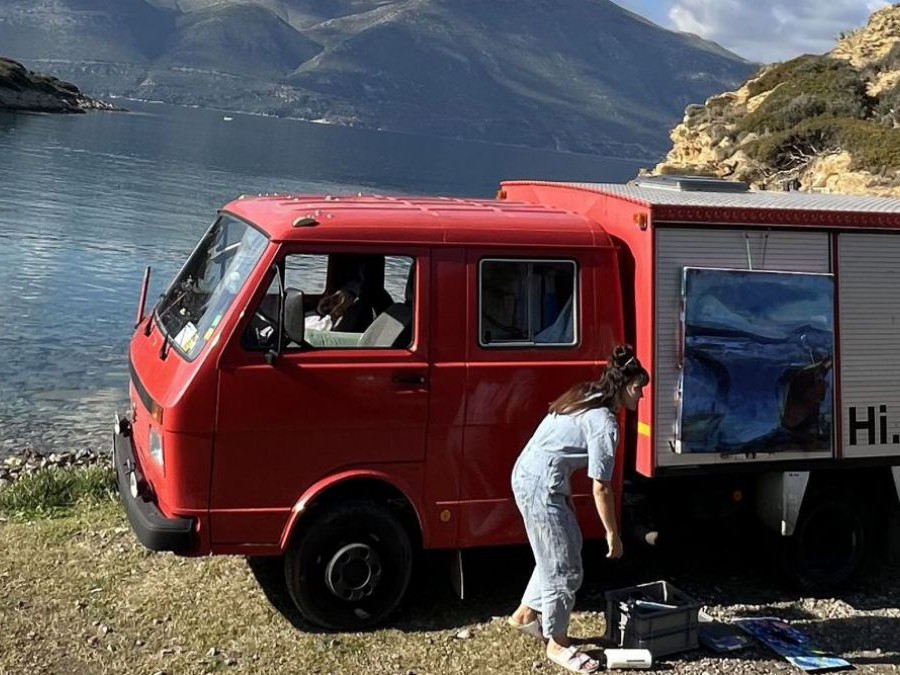 Am steinigen Ufer in einer Bucht parkt ein altes Feuerwehrauto, eine Frau im Overall bückt sich um Werkzeug aus einer Kiste. An der Ladewand des Fahrzeugs hängt eine bemalte, quadratische Leinwand.