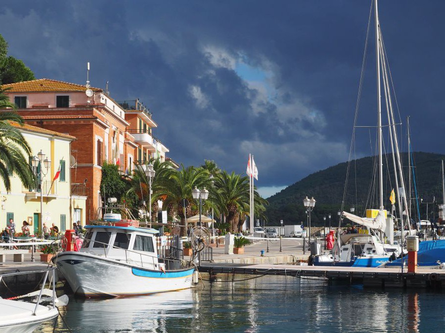 Sonnenbeschienener Hafen, Boote am Wasser, Prachtvolle Gebäude, Palmen, dunkle Wolken ziehen am Himmel auf.
