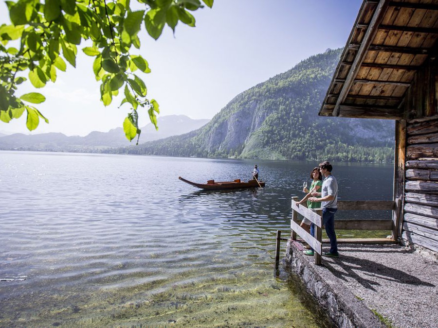 Vor einem Bootshaus steht ein Paar am Geländer und blickt auf den See, dessen Wasserfläche leicht gewellt ist. Jemand fährt mit einer Zille über den See. Im Hintergrund Gebirge, im Vordergrund hängt von oben ein leuchtend grün belaubter Zweig eines Baumes