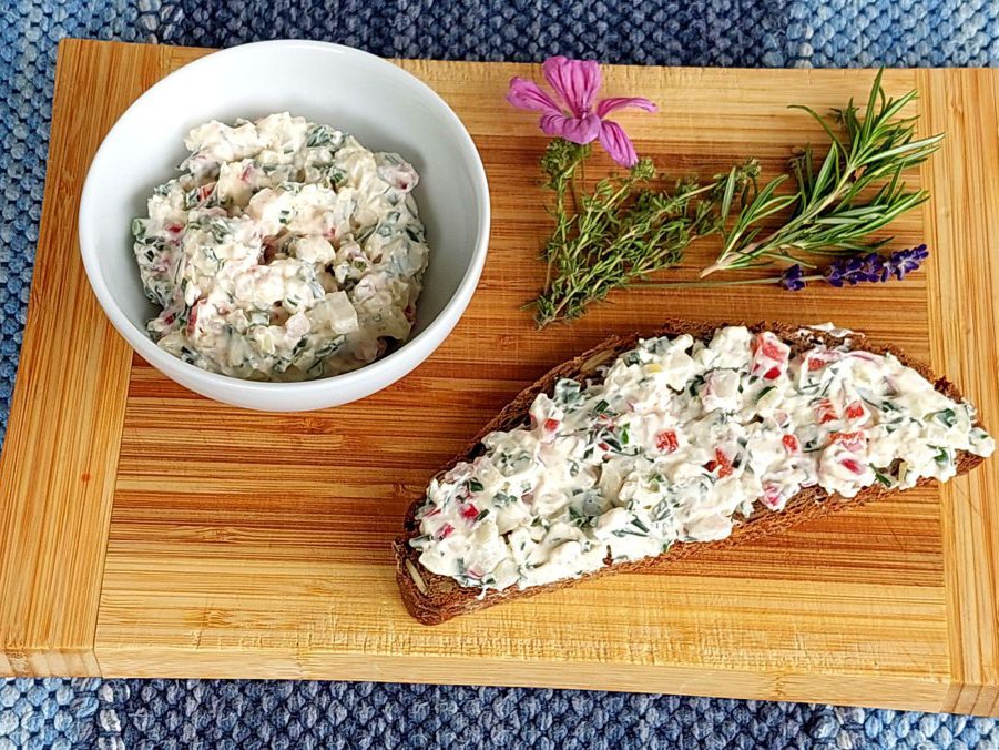 Auf einem Brett liegt ein Brot mit Kräuteraufstrich, daneben eine Schüssel mit diesem Aufstrich und einige Kräurer.