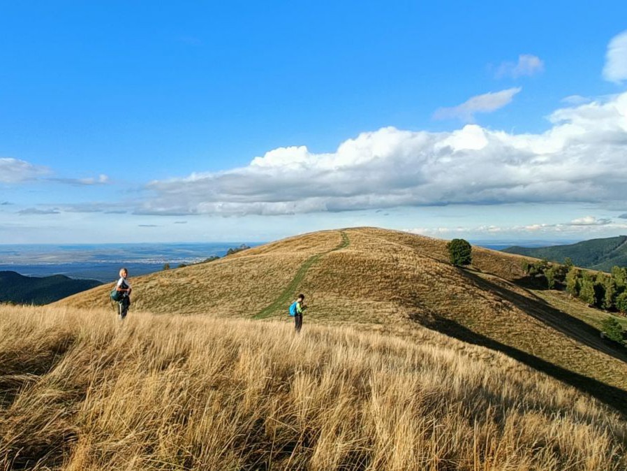 2 Personen auf einem hügeligen Wanderweg im hohen Gras. Extrem weite Fernsicht.