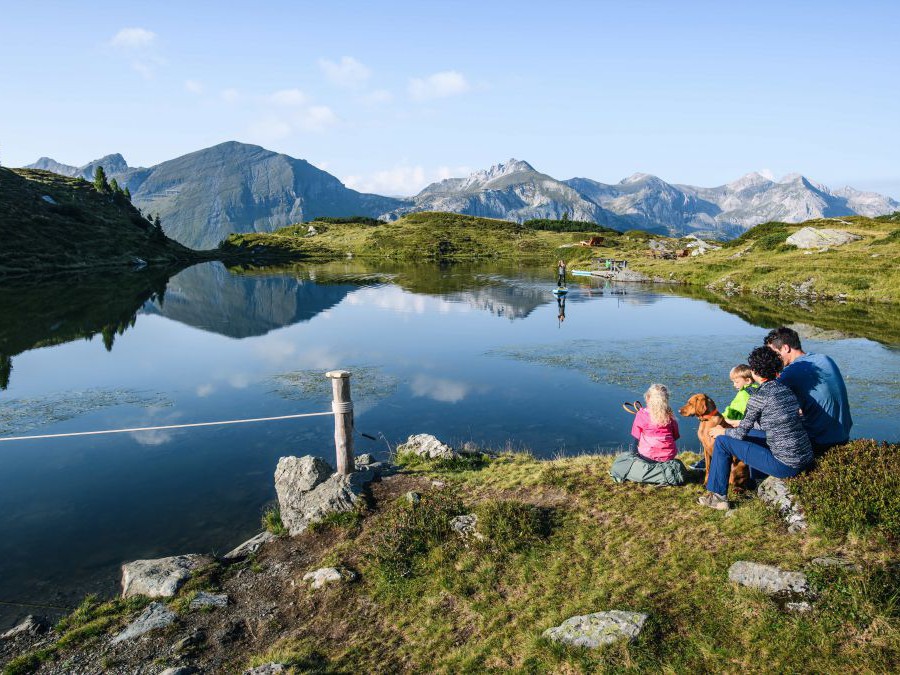 Familie mit 2 Kindern und Hund sitzt am Ufer eines Bergsees. Am See ein Stand Up Paddler. Im Hintergrund Bergpanorama.